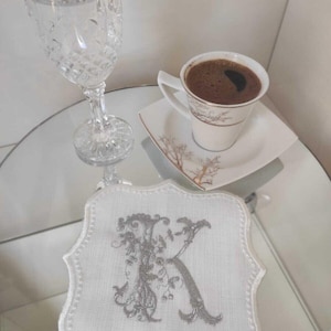 May include: A white embroidered coaster with a silver letter "K" and floral design sits on a mirrored surface. A cup of coffee on a saucer and a crystal water glass are also on the table.