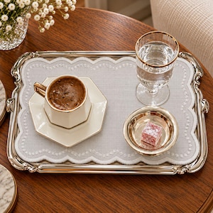 May include: A rectangular silver serving tray with scalloped edges holds a light grey fabric placemat with white embroidered trim. On the tray sits a hexagonal white coffee cup, a clear glass with a gold rim, and a small gold bowl with a pink cube.