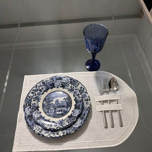 Pode incluir: Uma mesa posta com pratos com estampas florais azuis e brancas, uma taça de vinho azul e talheres em um sousplat branco. Os pratos têm uma cena de paisagem.