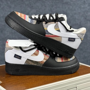 May include: Two Nike Air Force 1 sneakers. The shoes have a black base with a plaid pattern on the side panels and a white Nike swoosh. The soles are black, and the laces are black. The shoes are stacked on top of each other.