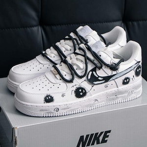 以下が含まれることがあります： 黒い落書き風のデザインと黒い靴紐が付いた、白いナイキエアフォース1スニーカー。スニーカーは灰色のナイキの箱の上にあります。箱にはナイキのロゴが見えます。