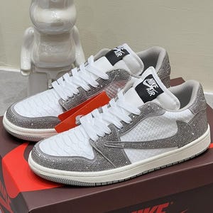 Pode incluir: Tênis branco e cinza de cano baixo com design texturizado e enfeites brilhantes. Os tênis têm cadarços brancos, gola cinza e o logotipo Nike Air na língua. Eles são exibidos em uma caixa de sapatos Nike marrom.