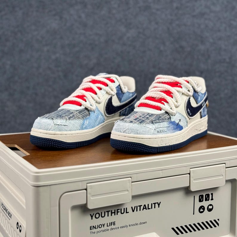 May include: A pair of white, blue, and red sneakers with a denim-like pattern. The shoes have a white sole with a navy blue outsole, red laces, and a navy blue Nike swoosh. The shoes are on top of a white storage container.