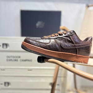 May include: Brown leather sneaker with a crocodile pattern. The shoe features a brown midsole and laces, with a Nike swoosh on the side. The sole has the letters "AIR" on the side.