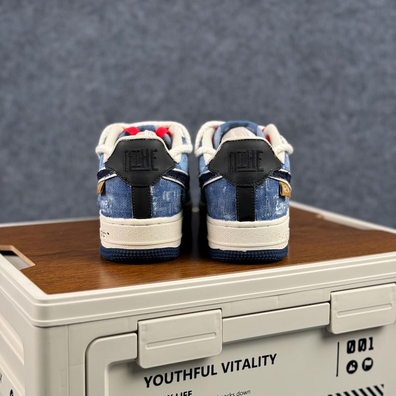 May include: A pair of blue denim sneakers with white soles and black accents. The sneakers have a white tongue and the word "NIKE" is visible on the back. The shoes are displayed on a wooden surface.