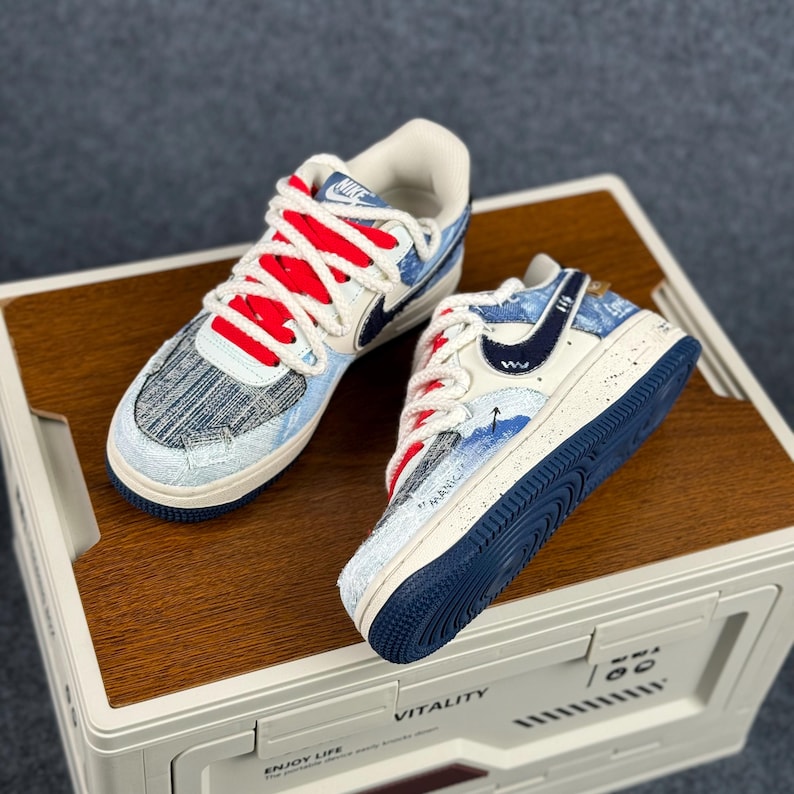 May include: Pair of Nike Air Force 1 sneakers. The shoes feature a white base with blue and denim-like accents, red laces, and a navy blue sole. The Nike swoosh is navy blue. The shoes are displayed on a wooden surface.