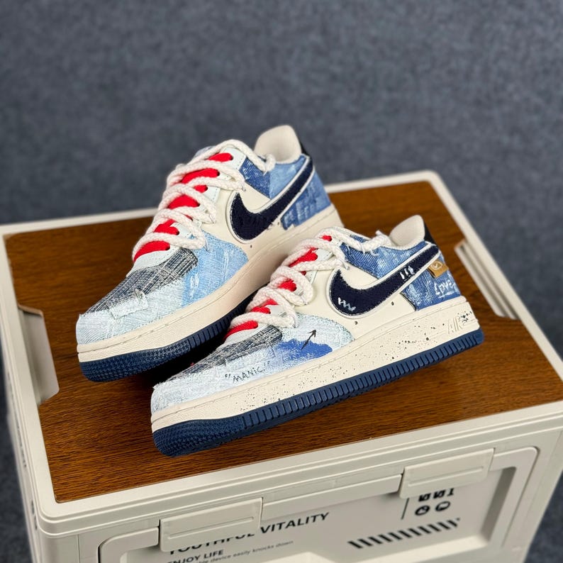 May include: Pair of Nike Air Force 1 sneakers with a denim-like design. The shoes feature a cream-colored base, navy blue accents, red laces, and the Nike swoosh logo. The text "MANIC" and "Love" are visible on the shoes.