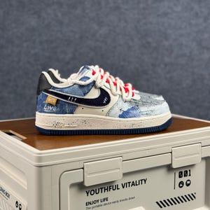May include: A blue and white Air Force 1 sneaker with a denim-like pattern. The shoe features a navy blue swoosh, red laces, and the word "Love" on the side. The sole is off-white with a navy blue trim.