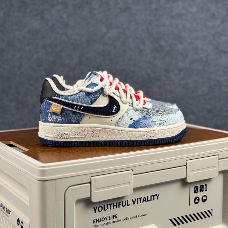 May include: A blue and white Air Force 1 sneaker with a denim-like pattern. The shoe features a navy blue swoosh, red laces, and the word "Love" on the side. The sole is off-white with a navy blue trim.