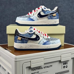 May include: Pair of blue and white sneakers with red laces. The shoes feature a denim-like pattern with navy blue accents, a white swoosh, and the word "Love" on the side. The soles are white with a navy blue trim.