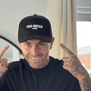 May include: A black baseball cap with the words "DAD SQUAD LONDON" in white lettering. The cap is being pointed at by a person. The person is smiling and pointing at the cap.