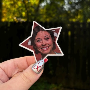 May include: A white star-shaped patch with a red background features a smiling woman with brown hair and a white shirt. The patch is being held by a hand with red nail polish and a silver clover design.