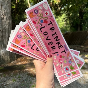 Może przedstawiać: Stos r&oacute;żowych naklejek z napisem "TRINKET LOVER!" i ilustracjami kot&oacute;w, wiśni i innych drobiazg&oacute;w. Naklejki są trzymane w dłoni na rozmytym zielonym tle.