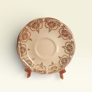 May include: A vintage saucer with a scalloped edge, featuring a cream-coloured centre and a decorative brown floral pattern. The rim is accented with gold trim. The saucer is displayed on a small, brown stand.