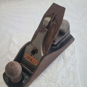 May include: A vintage Stanley hand plane, with a wooden handle and a metal blade. The plane has a dark brown body with the word "STANLEY" in orange lettering. The tool is designed for woodworking and shaping materials.