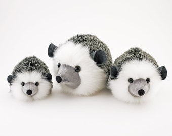 May include: Three plush hedgehogs with soft, white fur faces, gray noses, and black eyes. Their backs are covered in gray and white spiky material. The hedgehogs vary in size, with the largest in the center and the smallest on the left.
