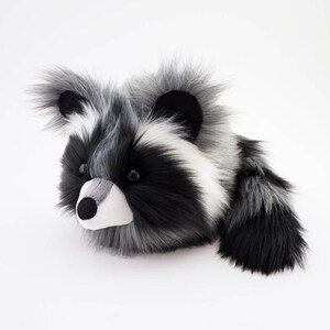 Pu&ograve; includere: Un giocattolo di peluche a forma di procione con pelliccia bianca e nera. Il procione ha il naso nero e gli occhi neri. Il giocattolo &egrave; sdraiato di lato con la coda arrotolata.