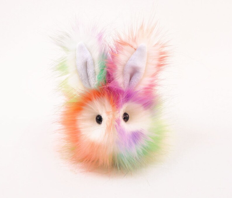 May include: A colourful fluffy toy bunny with white ears and black eyes. The bunny is made of soft, plush fabric and has a rainbow-coloured fur.