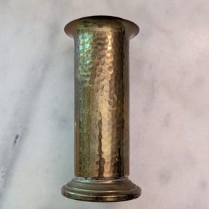 May include: A cylindrical brass vase with a hammered texture, featuring a flared rim and a circular base. A wooden ruler placed at the bottom shows the object stands approximately 4.5 inches tall and has a diameter of about 2 inches.