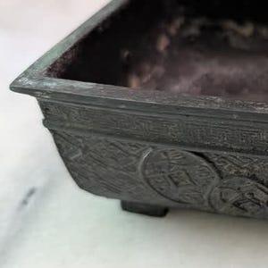 May include: A rectangular dark bronze-toned metal vessel with a wide rim and four small feet. The exterior features embossed circular coin motifs and a geometric border pattern along the top edge. The interior is deep, open, and dark in color.