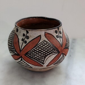 May include: A small, round, hand-painted clay pot featuring a cream-colored base with geometric patterns in terracotta orange and black. The design includes repeating floral-like shapes, cross-hatched squares, and thin black lines around the rim.