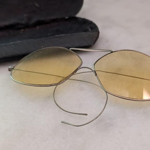 May include: Vintage-style eyeglasses featuring oval-shaped, light yellow tinted lenses set within a thin, metallic silver-toned wire frame. The glasses include distinctive coiled wire temples and are accompanied by a black, rectangular hard-shell case.
