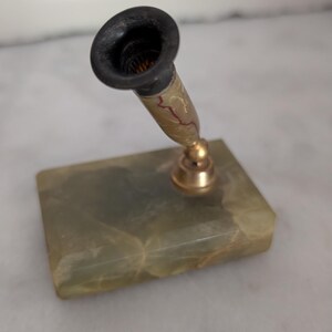 May include: A desk pen holder featuring a rectangular green stone base with a polished finish. An angled metal arm with a gold-toned finish connects to a tapered holder with a dark, matte-finished rim and a marbled, light-colored body.
