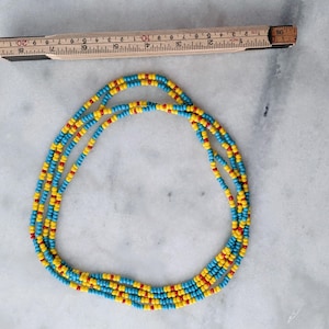 May include: A multi-strand necklace made of small, round beads in alternating colors of bright yellow, turquoise blue, and red. A wooden folding ruler is positioned above the necklace, showing a total length of approximately 20 inches.