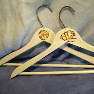 May include: Two wooden hangers painted light blue with a seashell and a fish design. The hangers are on a blue fabric surface.