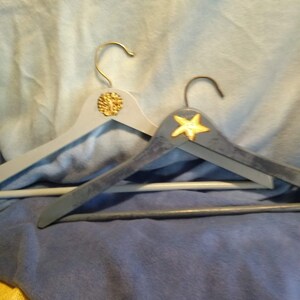 May include: Two wooden hangers painted in a light blue color. One hanger has a gold starfish design and the other has a gold seashell design.