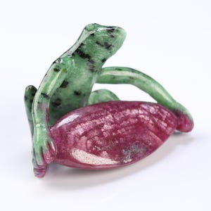 May include: A carved gemstone frog figurine in shades of green and black, cradling a deep red, oval-shaped stone. The frog is detailed with visible texture and sits upright, its arms wrapped around the red stone. The figurine is set against a white background.
