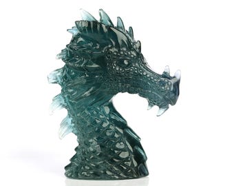 Puede incluir: Una escultura de cabeza de dragón translúcida de color verde azulado. La cabeza del dragón está detallada con escamas, cuernos y dientes afilados. La escultura está hecha de un material transparente, lo que permite el paso de la luz, creando un efecto brillante. La cabeza de dragón es un objeto decorativo.