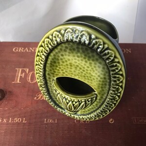 Peut inclure: Un vase en céramique vert de forme ovale avec un motif floral décoratif autour du bord. Le vase a une petite ouverture à l'avant pour insérer des fleurs.