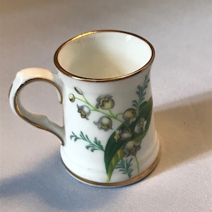Puede incluir: Una taza pequeña de cerámica blanca con un borde dorado y un diseño floral con flores de lirio de los valles y hojas verdes.