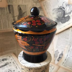 Peut inclure: Un récipient en bois peint en noir et or avec un couvercle. Le récipient présente un motif floral avec des baies rouges. Le récipient est posé sur un morceau de papier avec des notes de musique.