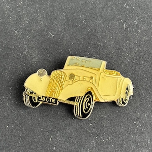 May include: A gold-colored vintage car pin with black tires and trim. The pin features the text "35 CTR" on the license plate. The car has a convertible top and is set against a dark background.