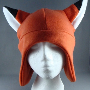 May include: An orange fox hat with black and white detailing. The hat is made of a soft fabric and has pointed ears. The hat is designed to cover the head and ears, with flaps that extend down the sides.