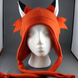 May include: A bright orange fleece fox hat with black and white ears and a long tail. The hat has a hood and is designed to fit over a person's head.