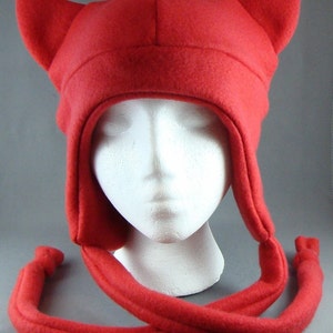 Puede incluir: Sombrero de forro polar rojo con cuernos, que se asemeja a un diablo, con una larga corbata de forro polar rojo. El sombrero está en una cabeza de maniquí blanca.