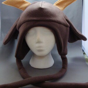 May include: A brown fleece hat with tan-colored ears and long, dangling ear flaps. The hat is designed to resemble a fictional character. The hat is displayed on a white mannequin head.