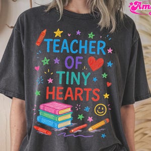 以下が含まれることがあります： 「TEACHER OF TINY HEARTS」の文字とカラフルなグラフィックがプリントされた濃いグレーのTシャツ。赤いハート、星、本、クレヨン、笑顔の顔がデザインされています。カジュアルで快適なスタイルです。