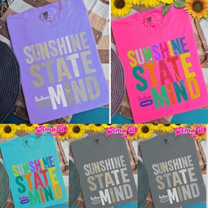 May include: Several t-shirts in shades of lavender, pink, teal, and grey. Each top displays the phrase "SUNSHINE STATE OF MIND" in a striking font. The shirts are presented flat, highlighting the design. The measurements are in centimeters.