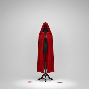 May include: A vibrant red hooded cape displayed on a black mannequin. The cape is made of a soft, flowing fabric and features a deep hood. The mannequin is on a black stand, set against a plain white background. Two small leaves are on the floor.