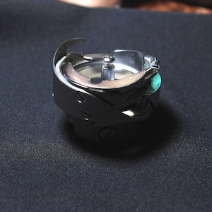 May include: A shiny, silver sewing machine bobbin case. The metal case has a curved hook and a central spindle. The bobbin case is on a dark, textured surface. Sewing machine parts.