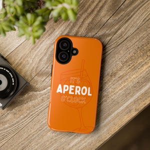 Könnte beinhalten: Eine leuchtend orangefarbene Smartphone-Hülle mit einer weißen Grafik eines Cocktailglases und dem Text IT'S APEROL O'CLOCK. Die Hülle verfügt über eine schwarze Aussparung für das Kameraobjektiv, passend für ein Gerät mit Doppelkamerasystem.
