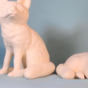 May include: Two white ceramic fox figurines. One fox is sitting with its head tilted up, while the other is lying down with its head resting on its paws.