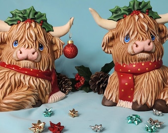 May include: Two brown Highland cow figurines wearing red and gold scarves and holly wreaths. Each cow has a Christmas ornament hanging from its ear. The ornaments are a red ball and a silver star. The cows are sitting on a light blue surface with gold and silver bows scattered around them.