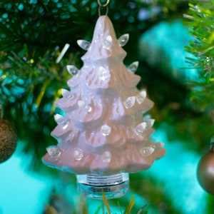 May include: A white ceramic Christmas tree ornament with small white lights. The ornament is hanging on a green Christmas tree.