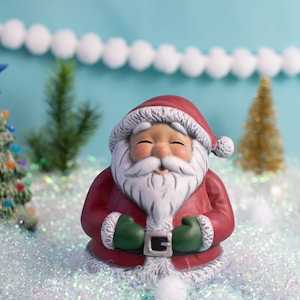 May include: A ceramic Santa Claus figurine with a red suit, white beard, and green gloves. A small Christmas tree with colorful lights and a blue star is in the background, along with a gold tree and a string of white pom-poms.