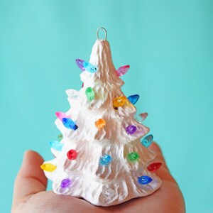 May include: A white ceramic Christmas tree ornament with colorful lights. The tree is decorated with multi-colored bulbs, creating a festive and whimsical look.